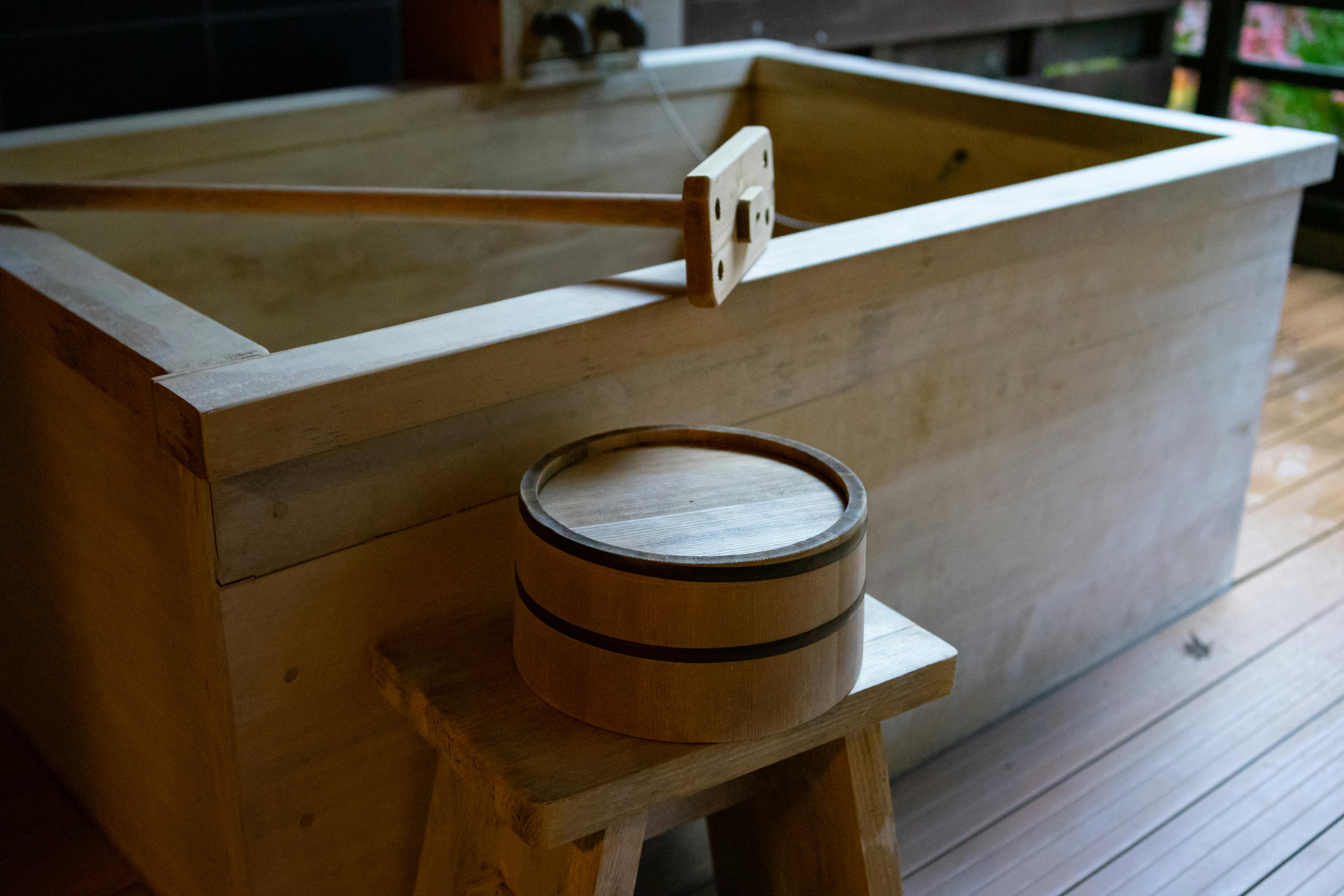 A traditional wooden Japanese bathtub sits beside a small wooden stool with a round wooden bucket on top, all set on wooden flooring in a serene, indoor setting.