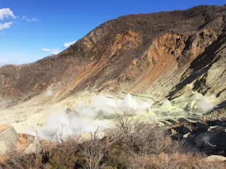 A rocky, barren hillside emits white steam from multiple vents, with sparse dry vegetation in the foreground and a clear blue sky above.