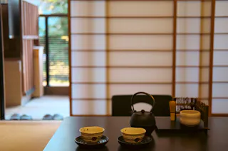 A Japanese tea set with two cups, a teapot, and utensils sits on a table in a traditional room with shoji screens and soft natural light coming from an open door.