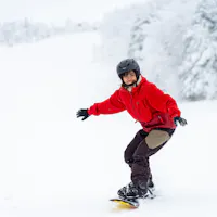 Snowboarding A person in a red jacket and black helmet is snowboarding down a snowy slope. The background is a winter landscape with snow-covered trees and distant figures on the slope. The person appears to be enjoying the ride.