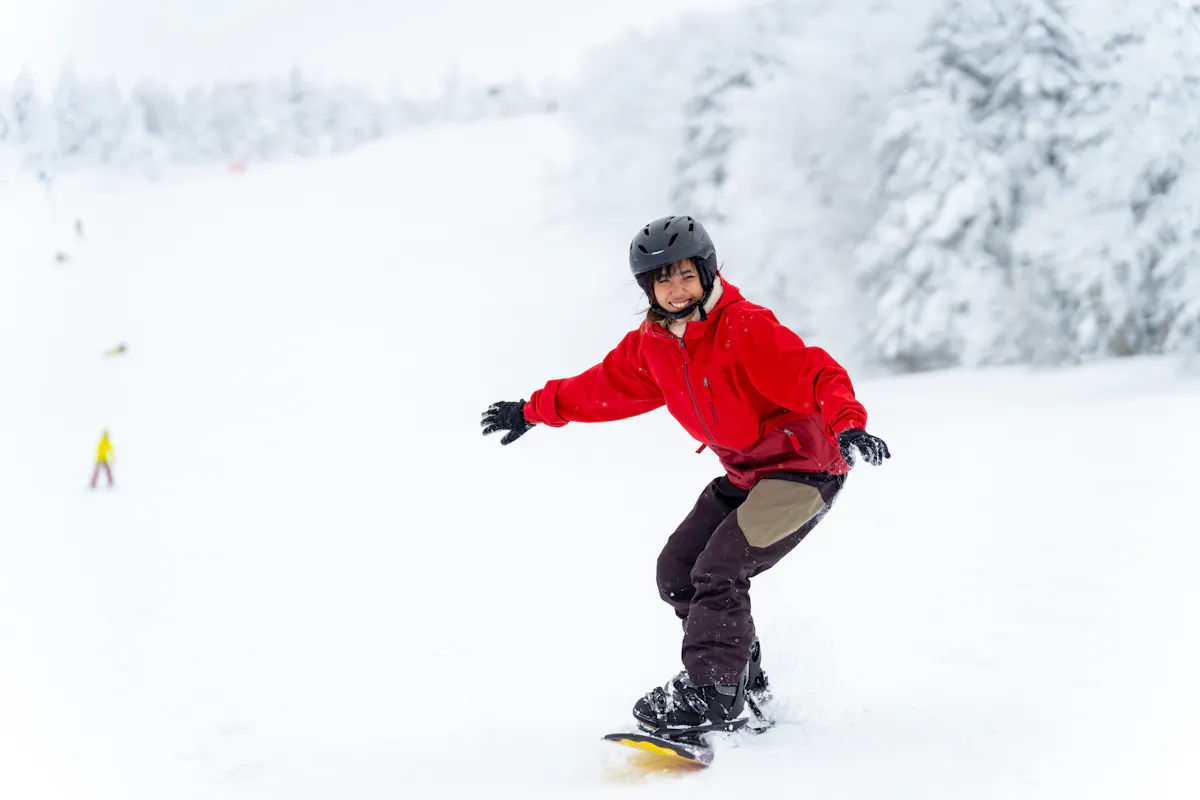 A person in a red jacket and black helmet is snowboarding down a snowy slope. The background is a winter landscape with snow-covered trees and distant figures on the slope. The person appears to be enjoying the ride.