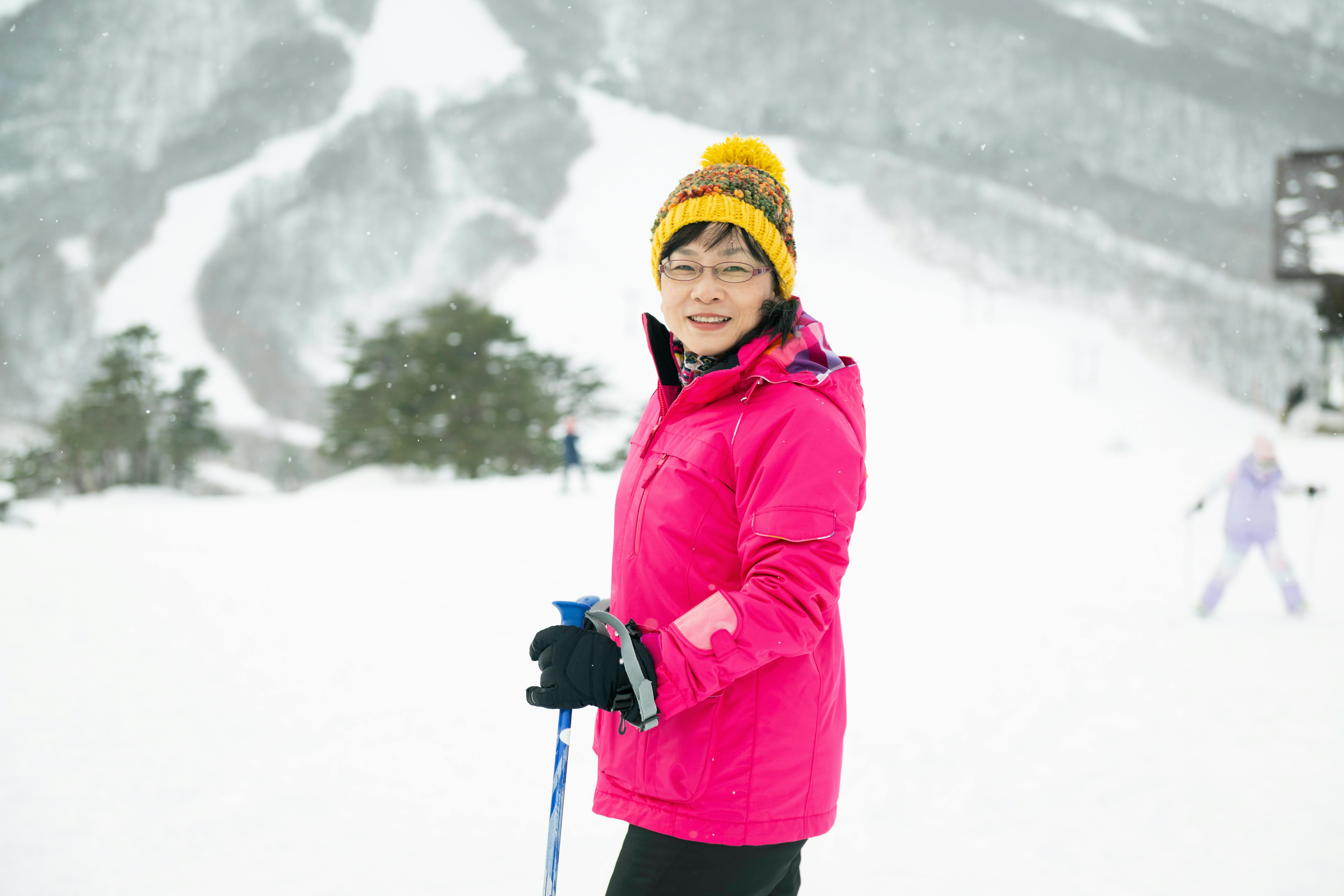 A woman in a bright pink jacket and yellow knit hat stands smiling in the snow, holding ski poles. Snowy mountains and trees are visible in the background, with a skier blurred in the distance.