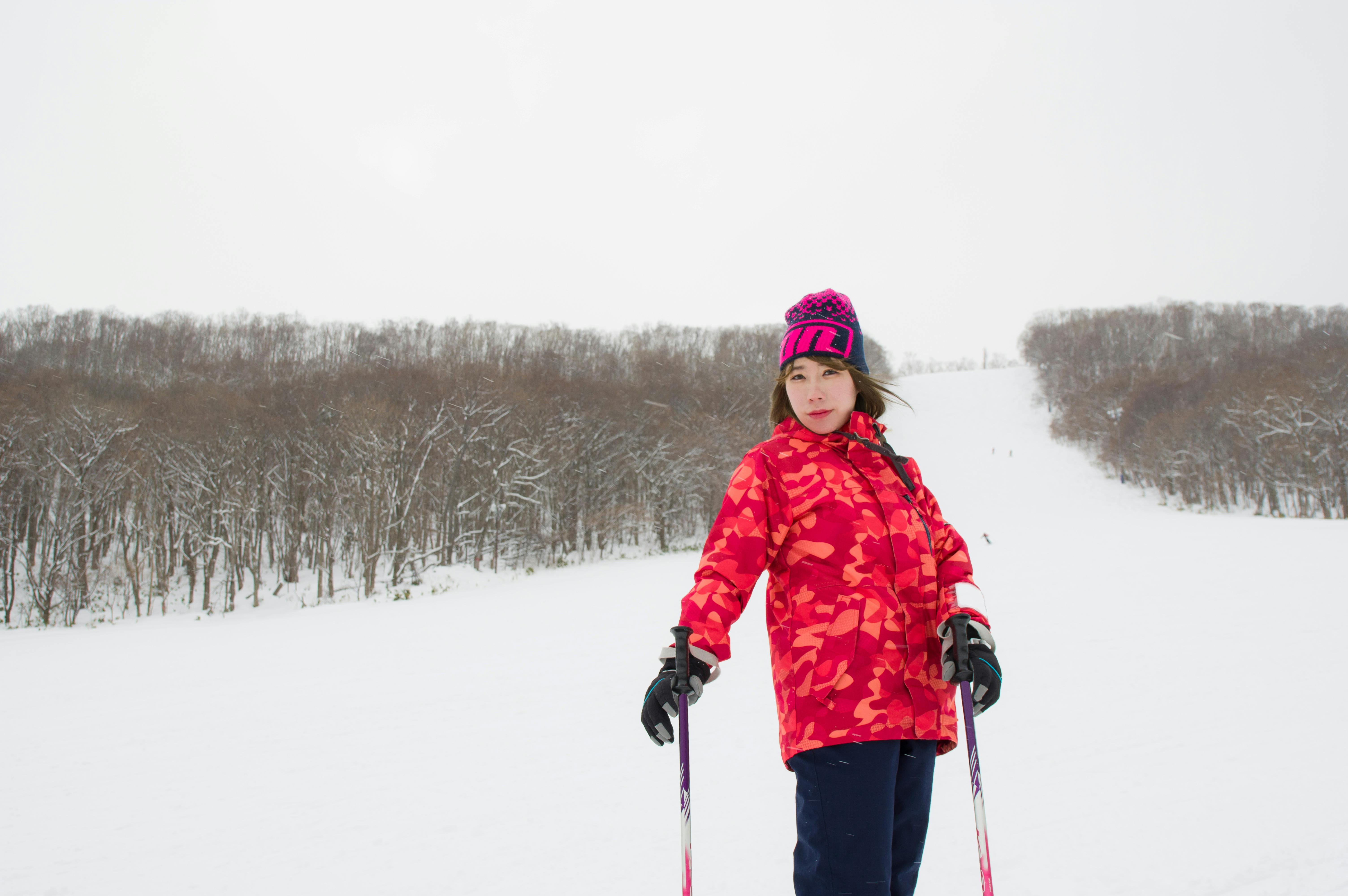 A young person in a bright pink patterned jacket and hat stands on a snowy slope with ski poles, with leafless trees and a hill in the background under a cloudy sky.