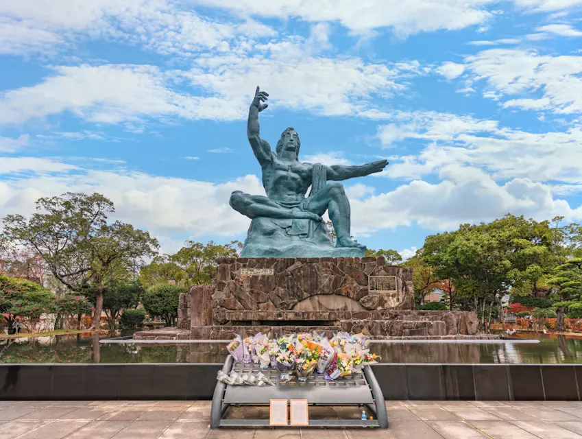 Peace Park The image depicts Nagasaki Peace Statue, a large bronze statue of a seated man with one arm pointing upward and the other outstretched to the side. The statue is surrounded by trees and a cloudy blue sky, with flowers laid at the base in tribute.