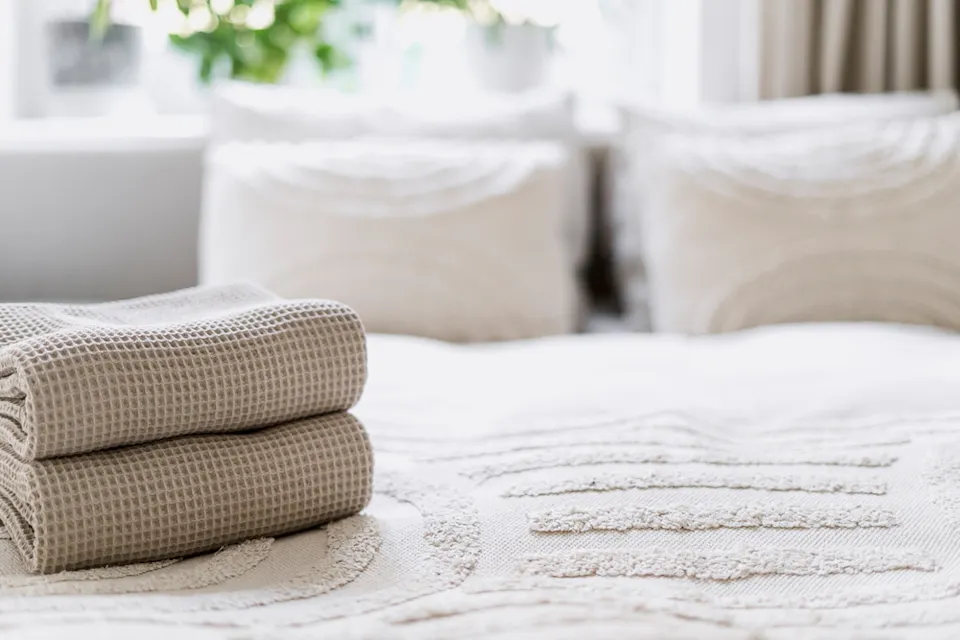 Hotel Room Two neatly folded, cream-colored waffle weave towels are placed on a soft, textured bedspread in a bright, airy bedroom. The background includes two plush, patterned pillows, a window with natural light, and green plants.