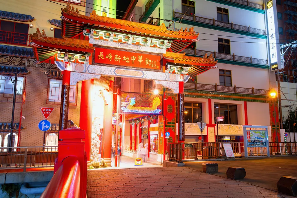 Nagasaki Shinchi Chinatown A vibrant, ornate entrance gate to a Chinatown area is illuminated at night. It showcases traditional Chinese architectural details with red columns, intricate carvings, and colorful decorations. Buildings surround the gate, including a map and signposts nearby.