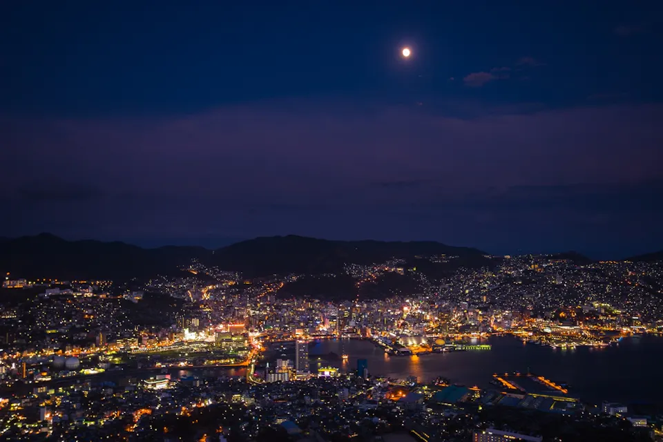 Night Views from Mount Inasa A nighttime aerial view of a city with a full moon in the sky. The city lights illuminate the urban landscape, including tall buildings, roads, and a waterfront area. Hills surround the city, and the dark blue night sky contrasts with the bright city lights.