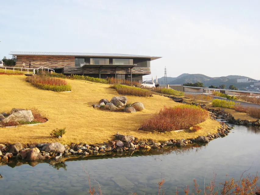 Garden Terrace Nagasaki Hotel A modern building with a flat roof and large windows is perched on a hill overlooking a landscaped area with a pond. The landscape features tiers of golden grass, shrubs, and rocks, with a backdrop of distant mountains under a clear sky.
