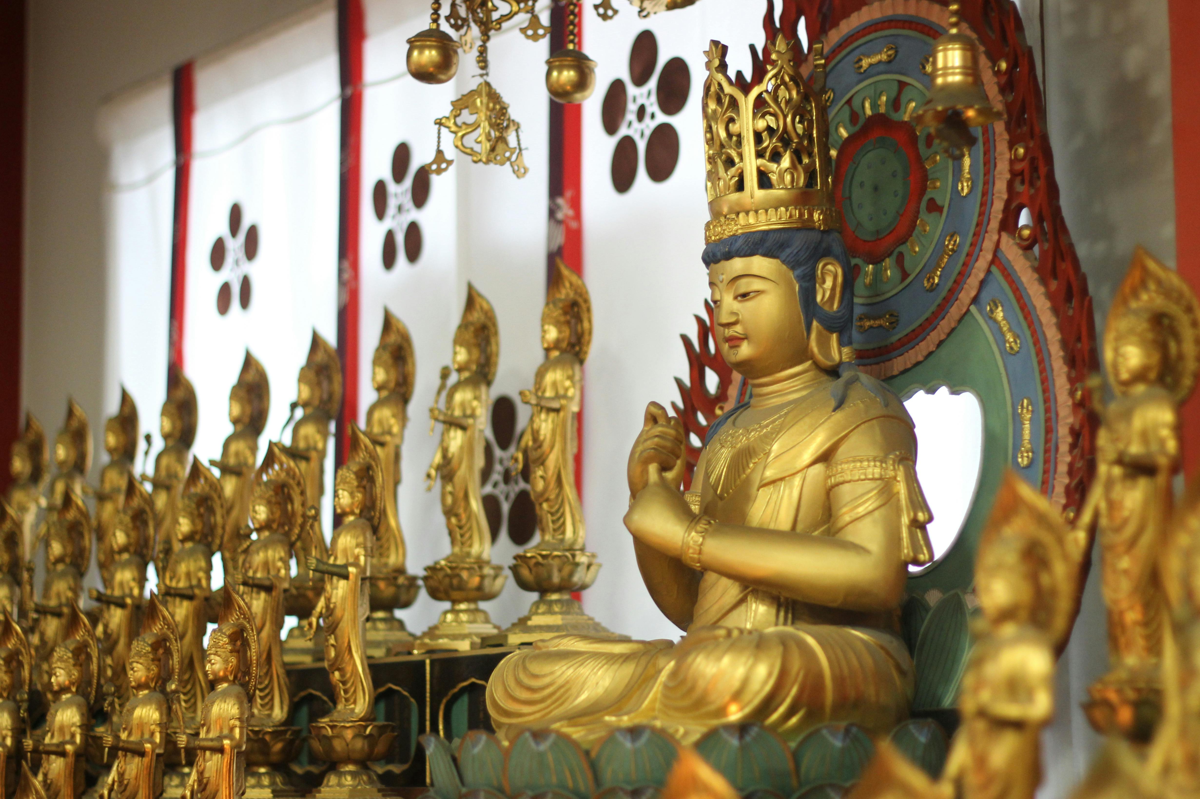 Golden Buddhist statues, including a central large figure wearing a crown and ornate attire, are arranged on an altar. Smaller statues surround the central figure, and decorative elements can be seen in the background.