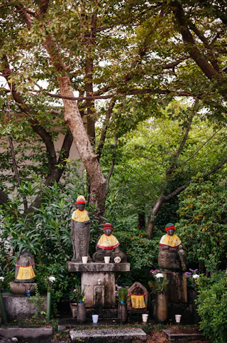 Four stone statues wearing red hats and yellow bibs sit on stone platforms, surrounded by lush green trees and plants. Flowers and small cups are placed at the statues’ bases as offerings.