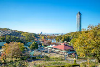 A scenic park with green trees, walking paths, and a red-roofed building, with a tall glass tower and cityscape in the background under a clear blue sky.