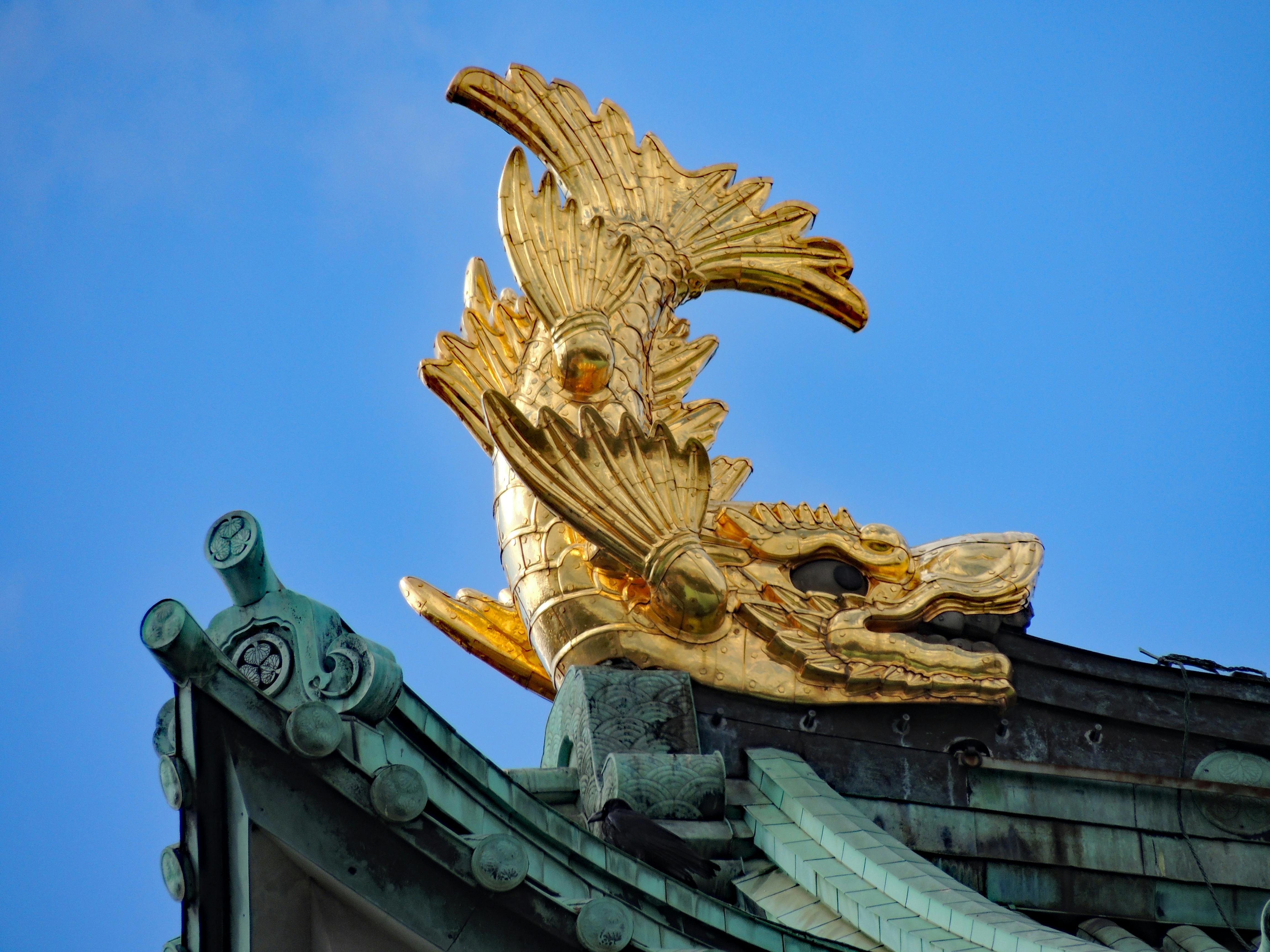 A close-up of a golden shachihoko, a mythical fish with a tiger’s head, perched on the green-tiled roof edge of a traditional Japanese building, set against a clear blue sky.