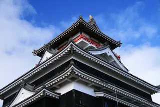 A close-up view of a traditional Japanese castle with tiered roofs, ornate eaves, and red accents, set against a bright blue sky with scattered clouds.