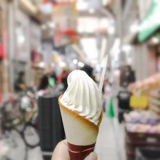 A hand holds a vanilla soft-serve ice cream cone with a spoon, against the blurred background of a busy indoor market or shopping arcade.