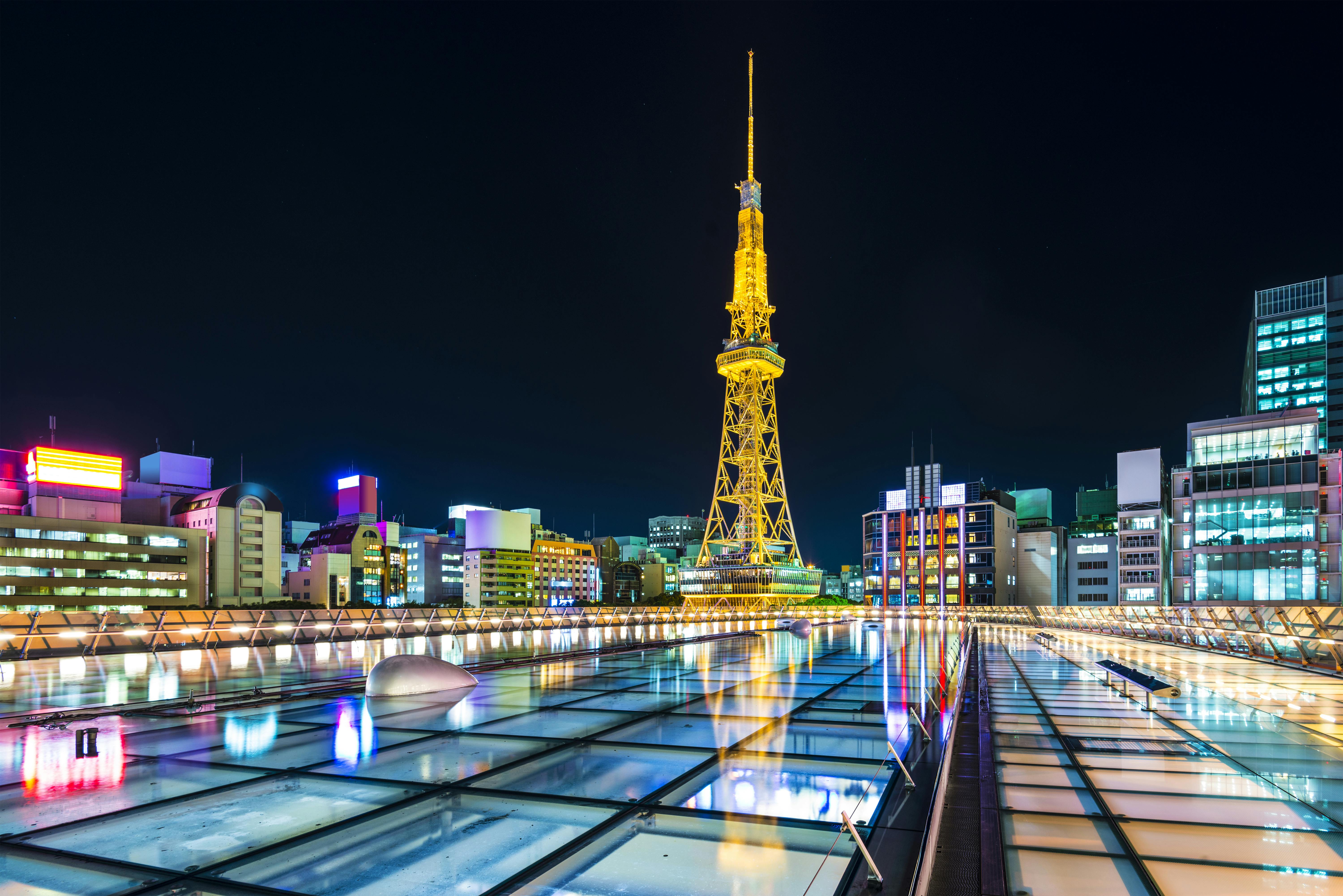 Night view of Nagoya TV Tower illuminated in yellow, surrounded by city buildings with colorful lights, reflected on the glass roof of Oasis 21 in Nagoya, Japan.