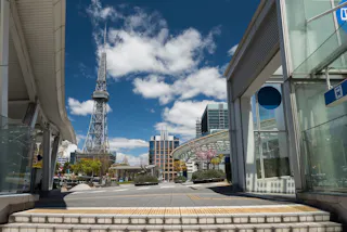 A modern urban plaza under a blue sky with scattered clouds, featuring a tall steel tower, contemporary buildings, glass structures, and trees with some pink blossoms.