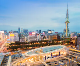 A cityscape at dusk shows modern buildings, busy streets with light trails from cars, a glass-roofed structure in the foreground, and a tall tower with an observation deck on the right. The sky is clear and blue.