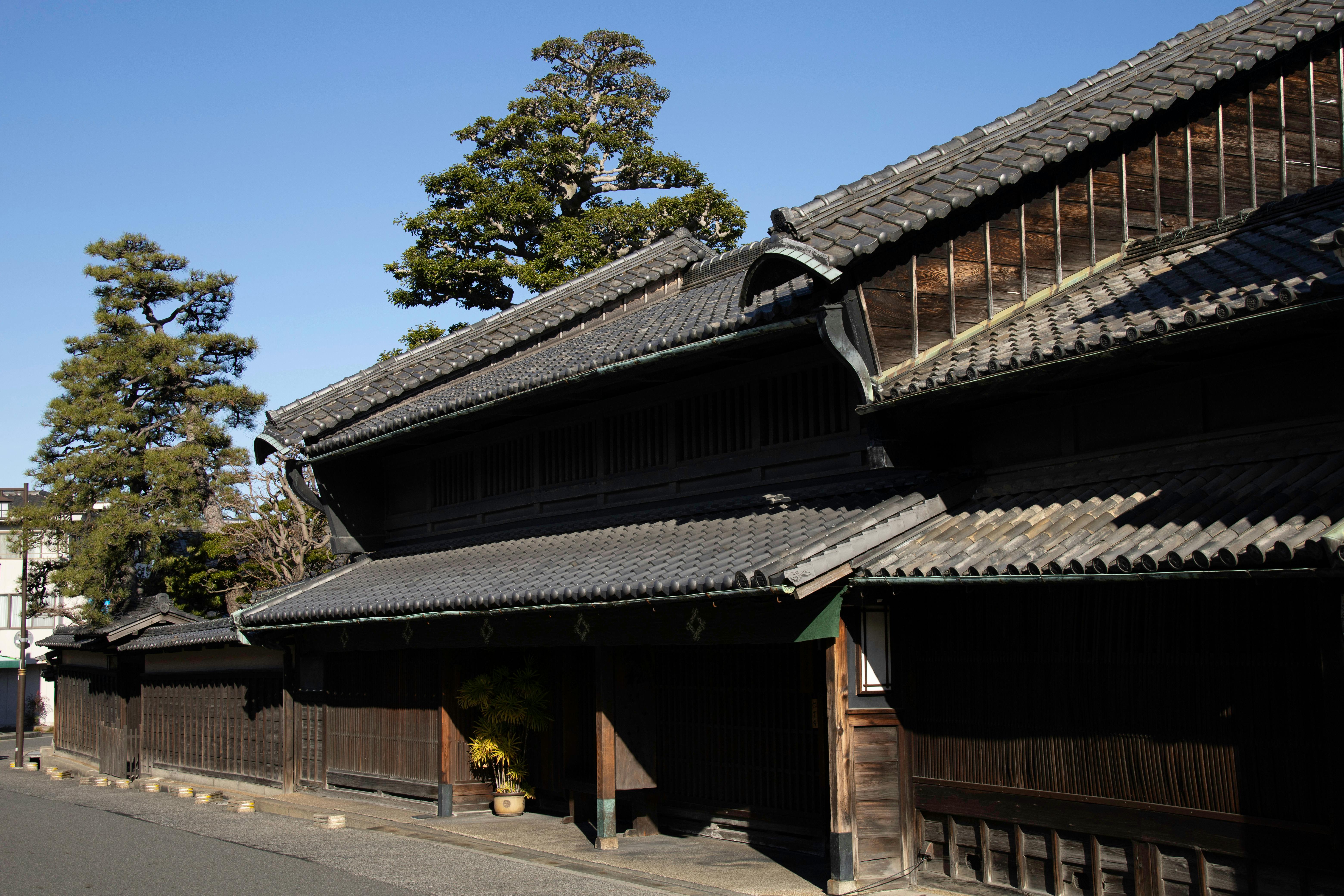A traditional Japanese wooden building with a tiled roof stands in sunlight on a quiet street, surrounded by tall green trees under a clear blue sky.