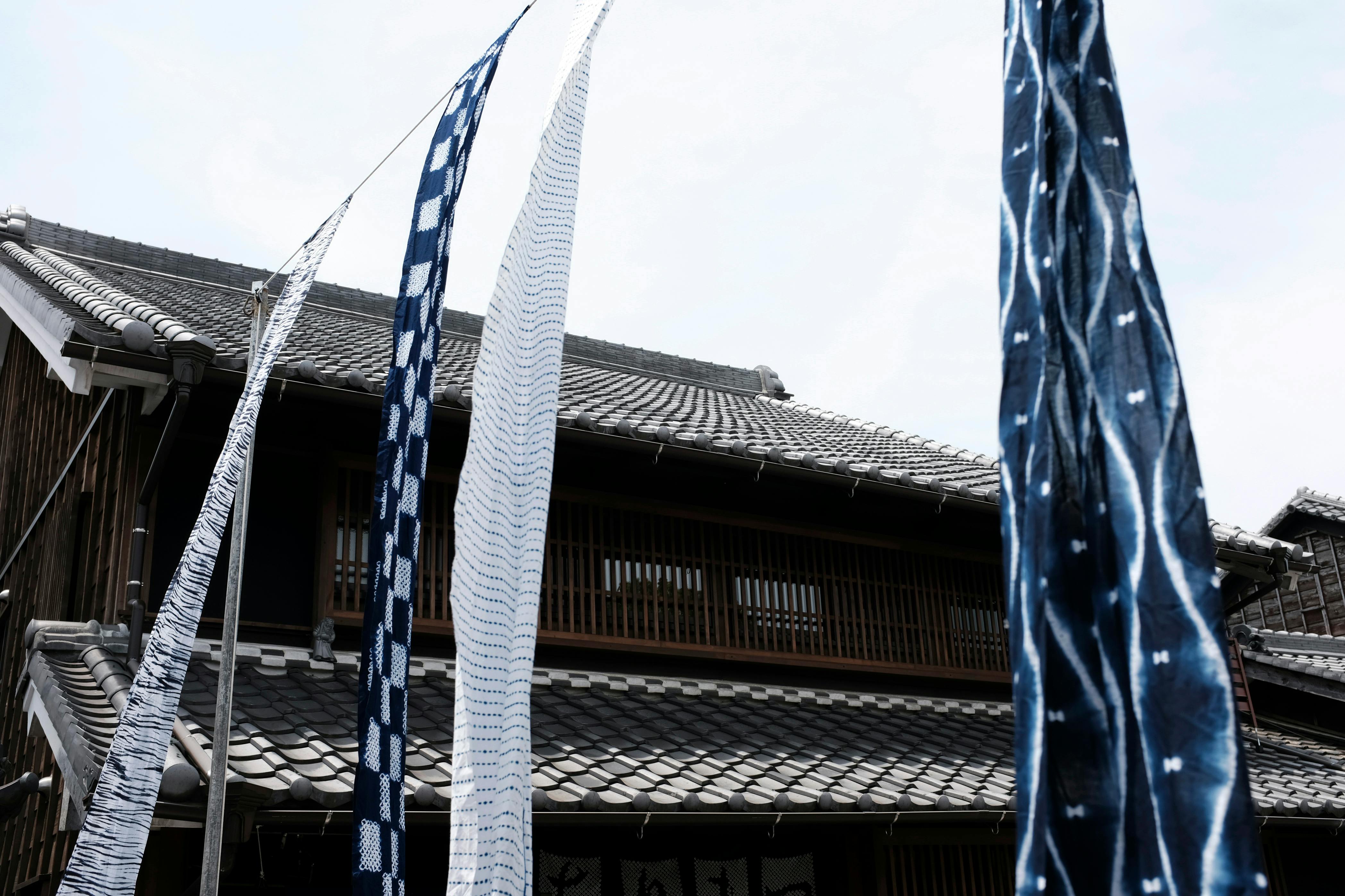 Traditional Japanese wooden building with a tiled roof, viewed from below, with long indigo and white patterned fabric banners hanging in the foreground against a bright sky.