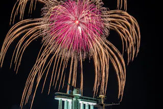 A large pink and gold firework bursts in the night sky above a brightly lit building with columns and antennas, creating a dramatic and festive scene.