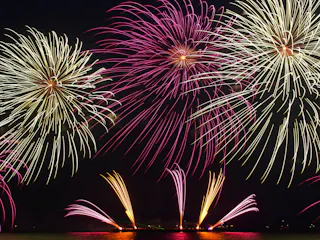 Colorful fireworks burst in the night sky above a body of water, reflecting vibrant yellow and pink light. Multiple large and small fireworks are visible, creating a festive and celebratory atmosphere.
