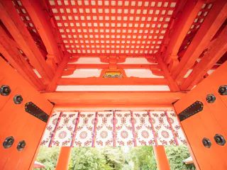 View from beneath a traditional Japanese torii gate, showcasing its bright red beams, patterned ceiling, and decorative hanging curtain, with greenery visible outside.