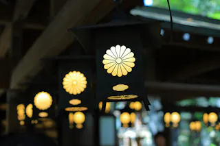 Black metal lanterns with glowing lights hang from a wooden ceiling. The lanterns feature a cutout chrysanthemum flower design, casting warm light in a shaded area with soft greenery visible in the background.