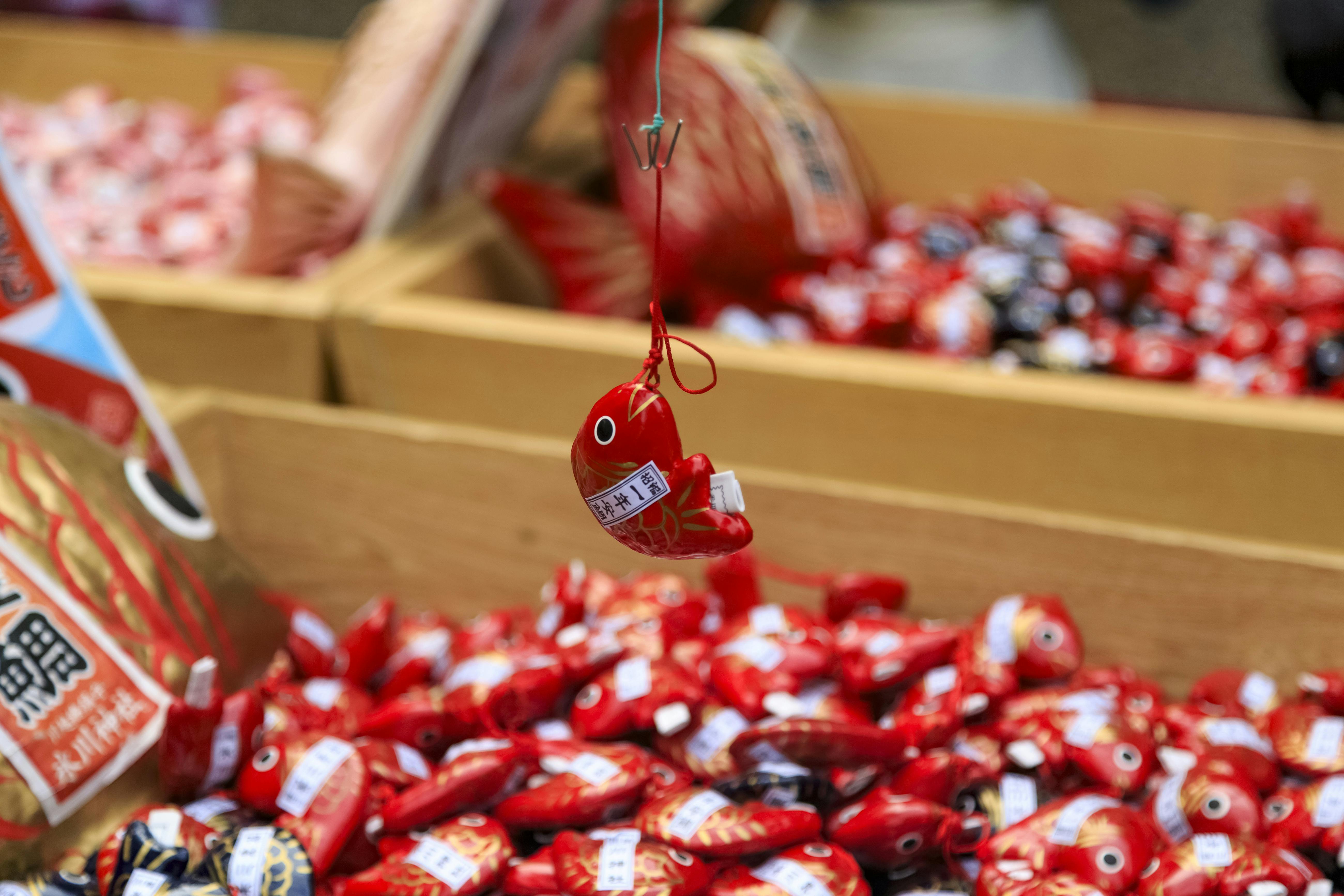 A small red fish-shaped charm, decorated with Japanese writing, hangs by a string above a box filled with similar red and black fish charms at a market or festival stall.