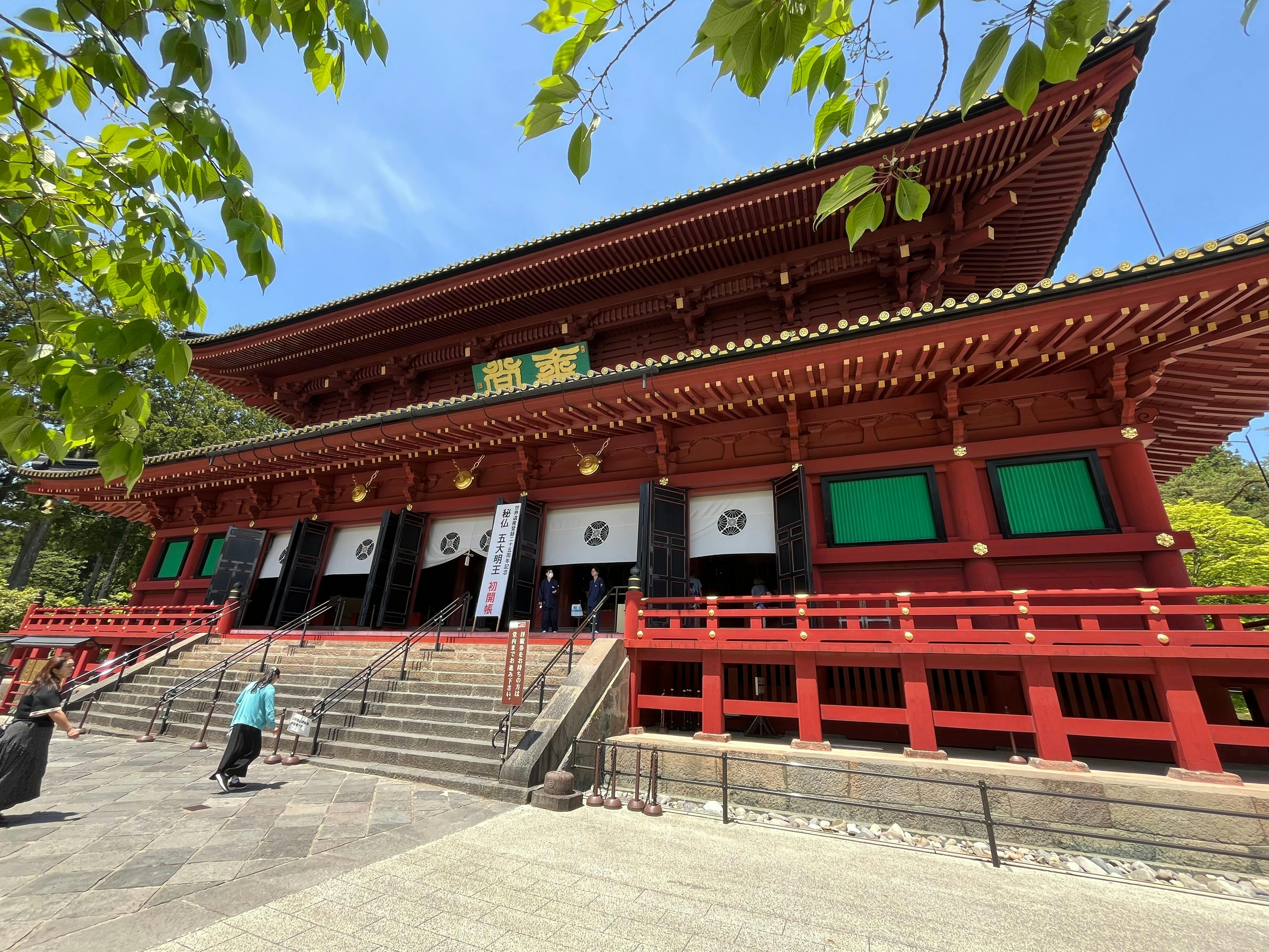 A large traditional Japanese temple with bright red wooden beams, gold accents, and ornate architectural details, surrounded by greenery. People walk up the steps toward the entrance under a sunny sky.