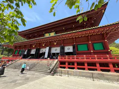 A large traditional Japanese temple with bright red wooden beams, gold accents, and ornate architectural details, surrounded by greenery. People walk up the steps toward the entrance under a sunny sky.