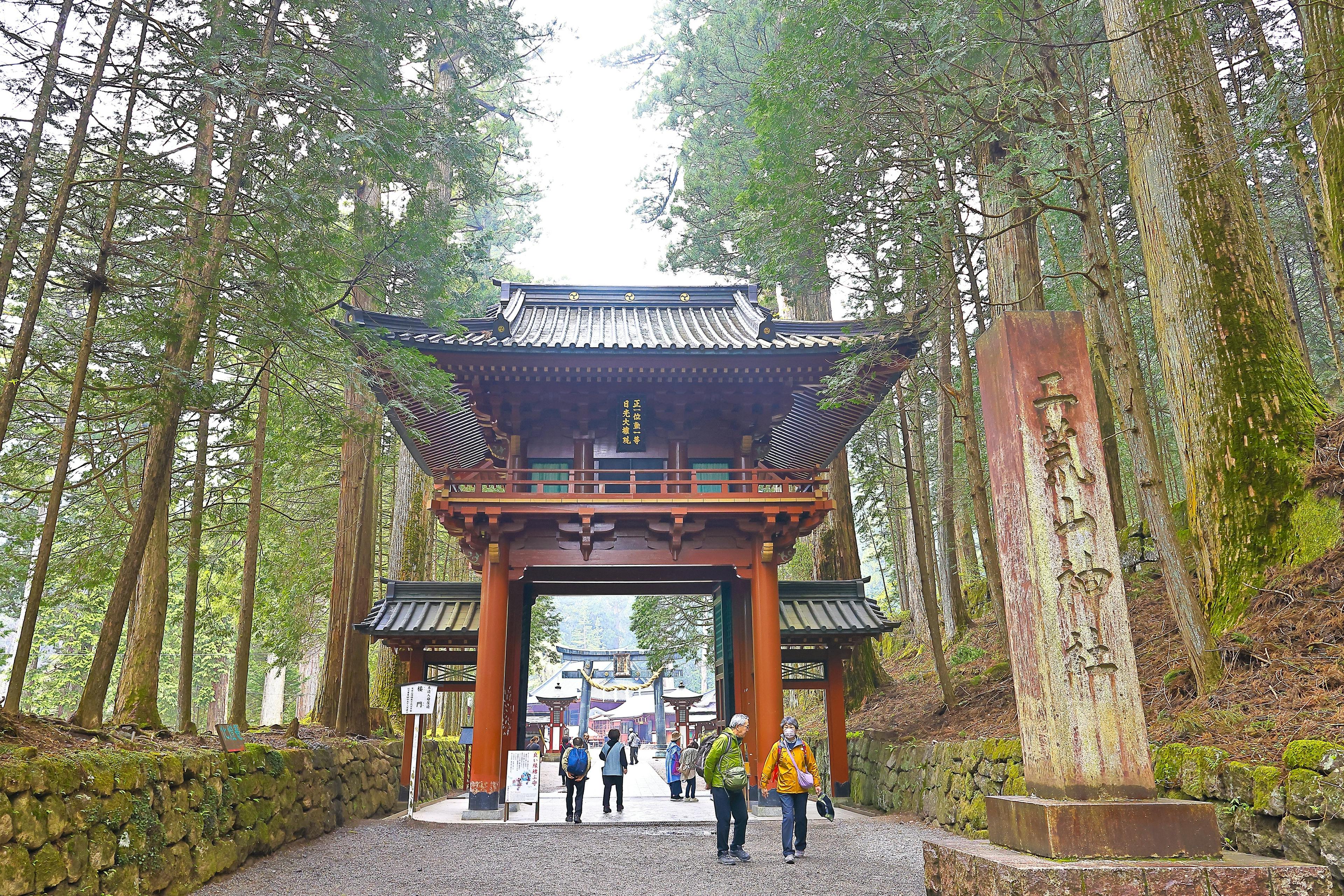 Visitors walk through a traditional Japanese wooden gate surrounded by tall trees, entering a temple complex. A large stone monument with inscriptions stands to the right of the path.