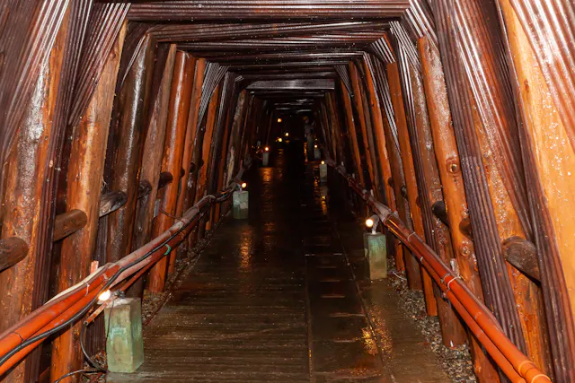 Dimly lit tunnel with wooden beams supporting the walls and ceiling. The pathway is wet, with orange pipes and small lights running along both sides, creating a sense of depth and perspective.