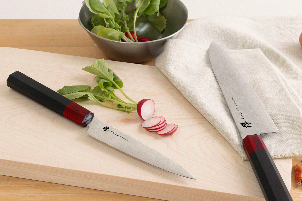 Taski Knives Two knives are resting on a wooden surface next to some sliced radishes and a metal bowl filled with leafy greens. One knife is laying flat with the blade facing up, and the other is partially sheathed. A white cloth is also visible in the background.