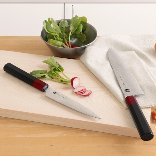 Taski Knives Two kitchen knives with Japanese inscriptions are placed on a wooden surface, accompanied by sliced radishes and a metal bowl containing radish greens. A white cloth lies under one of the knives, and the background features a light-colored cabinet.