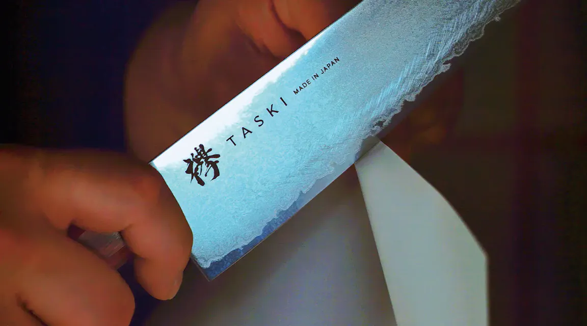 A close-up of a hand carefully holding a Japanese chef's knife, with the brand name "TASKI" and "Made in Japan" engraved on the blade. The knife is slicing through a piece of white paper, demonstrating its sharpness.