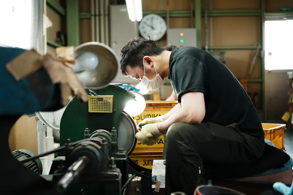 A man wearing a mask and gloves operates a machine in a workshop. He is focused, sitting next to several labeled containers and under fluorescent lights, surrounded by various tools and materials. A clock is visible on the wall in the background.