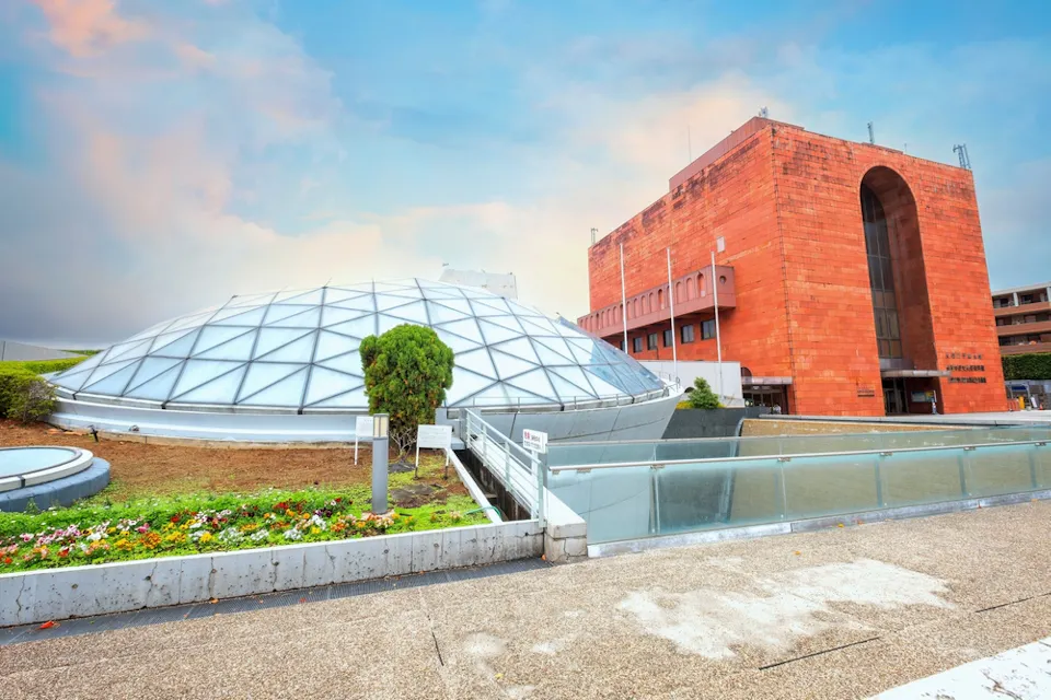 Nagasaki Atomic Bomb Museum The image shows a modern building with a unique, dome-shaped glass roof next to a red brick structure with large, arched entrance. The foreground features a concrete walkway with garden and railing. The sky is partly cloudy with a hint of warm evening light.