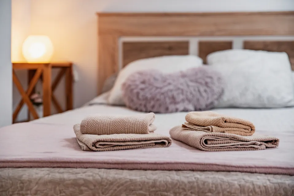 Hotel Room A neatly made bed with a wooden headboard is adorned with white and textured gray pillows, including a fluffy heart-shaped pillow in the center. Two neatly folded beige towels are placed on the foot of the bed. A lit lamp on a wooden side table adds a warm glow.