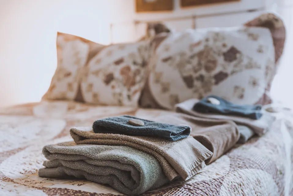 Hotel Room Folded towels and washcloths are neatly arranged on a bed adorned with a patterned quilt and pillows in a cozy, softly lit room. The bedding features floral designs, adding to the warm and inviting atmosphere.