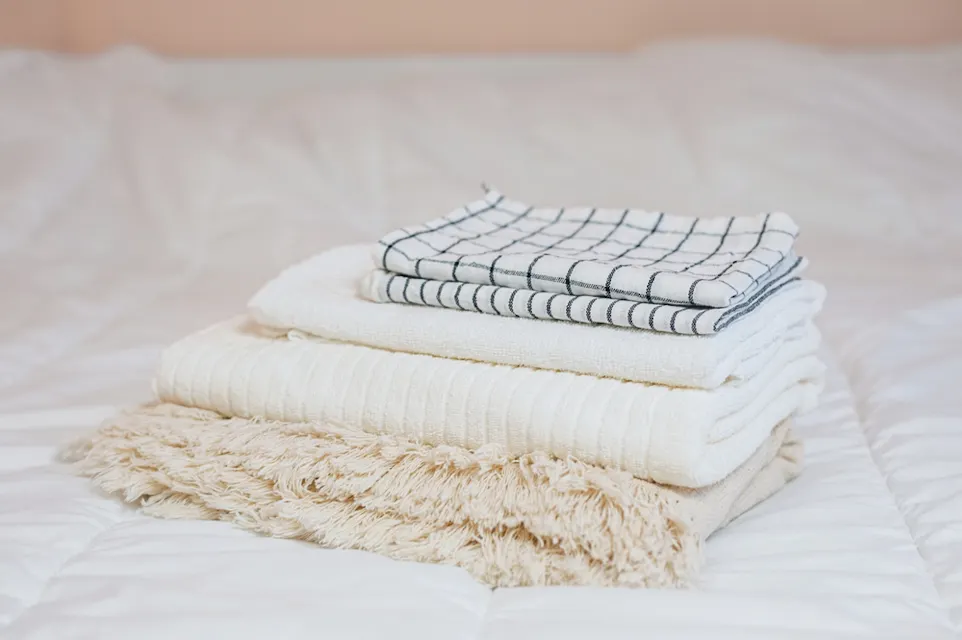 Hotel Room A neatly folded stack of towels is seen on a bed. The stack includes a checkered towel on top, followed by a white towel, a ribbed cream-colored towel, and a beige towel with a soft, fluffy texture. The bed has a white quilted cover.