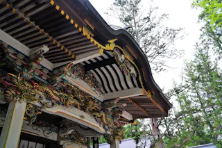 Ornate roof of a traditional Japanese temple with intricate wooden carvings and colorful decorative details, set against tall green trees and a bright sky.