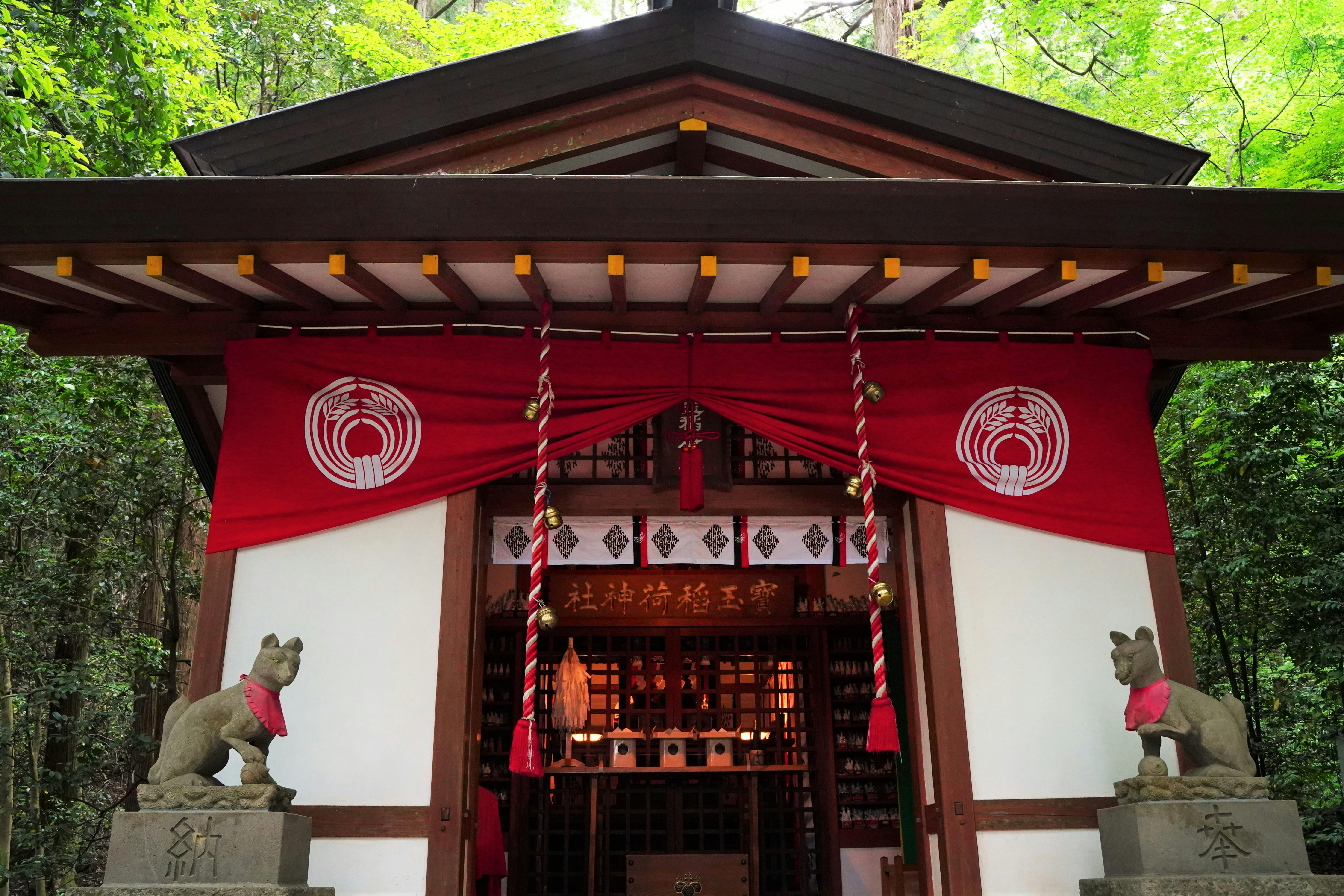 A small Japanese shrine surrounded by lush greenery, with two fox statues wearing red bibs at the entrance and red banners hanging from the roof. The shrine features wooden details and decorative ropes.