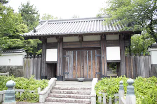 A traditional Japanese wooden gate with a tiled roof stands at the top of stone steps, surrounded by lush green trees and foliage. The gate is part of a fence with stone walls on either side.