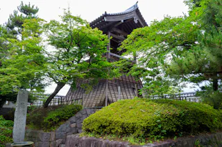 A traditional Japanese wooden bell tower stands among lush green trees and shrubs, with a stone marker and rock-lined landscaping in the foreground. The structure features a tiled roof and intricate wooden details.