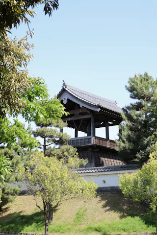 A traditional Japanese wooden bell tower with a tiled roof stands surrounded by green trees and shrubs under a clear blue sky.