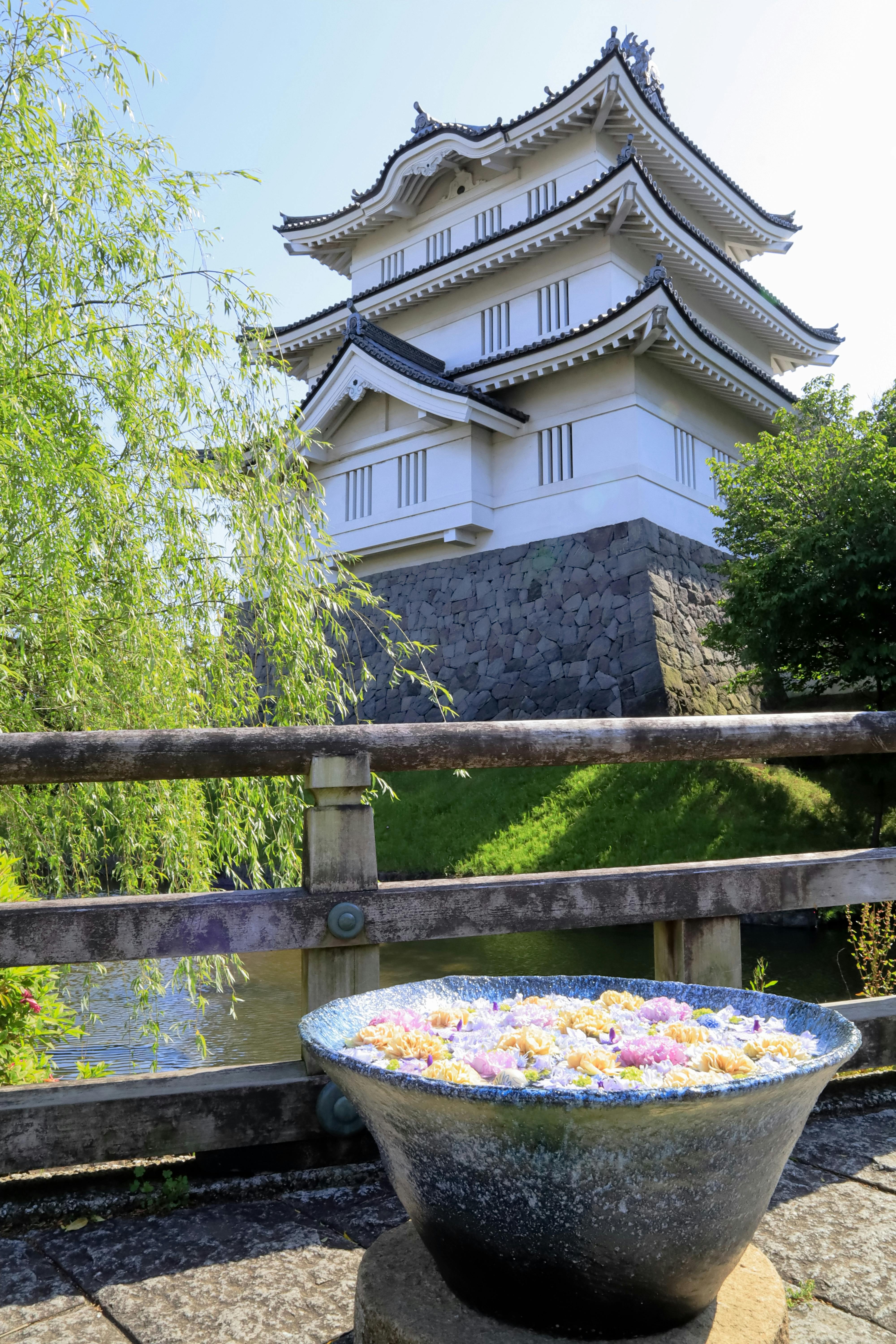 A traditional Japanese castle with white walls and tiled roofs stands behind a wooden bridge and a large bowl filled with floating colorful flowers on a sunny day.