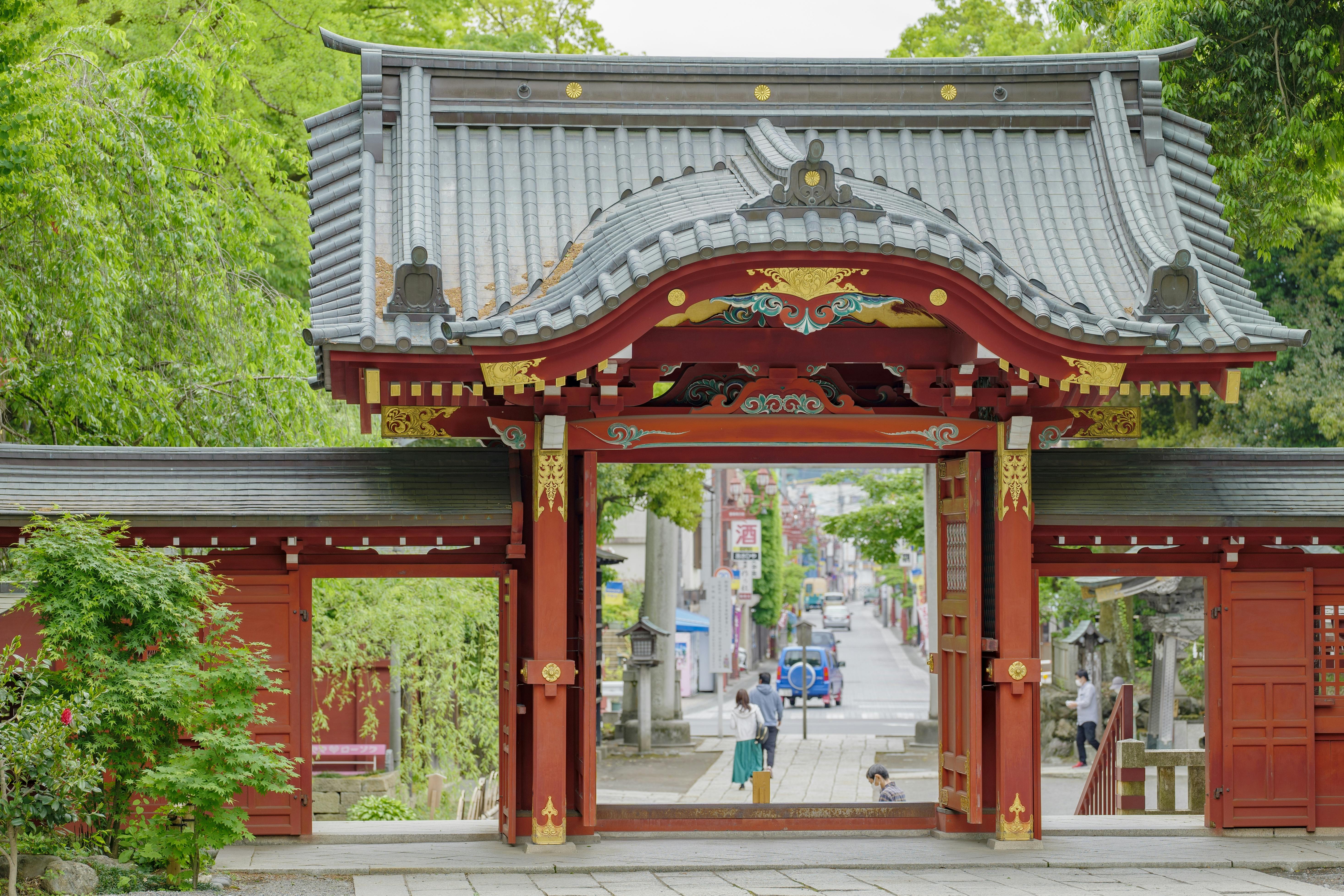 A traditional Japanese temple gate with ornate roof and red wooden pillars frames a view of a tree-lined street and people walking in the background. Lush greenery surrounds the structure.