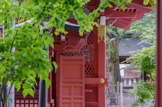 A close-up of a traditional red Japanese shrine partially obscured by green leaves, with intricate architectural details and a peaceful outdoor setting.