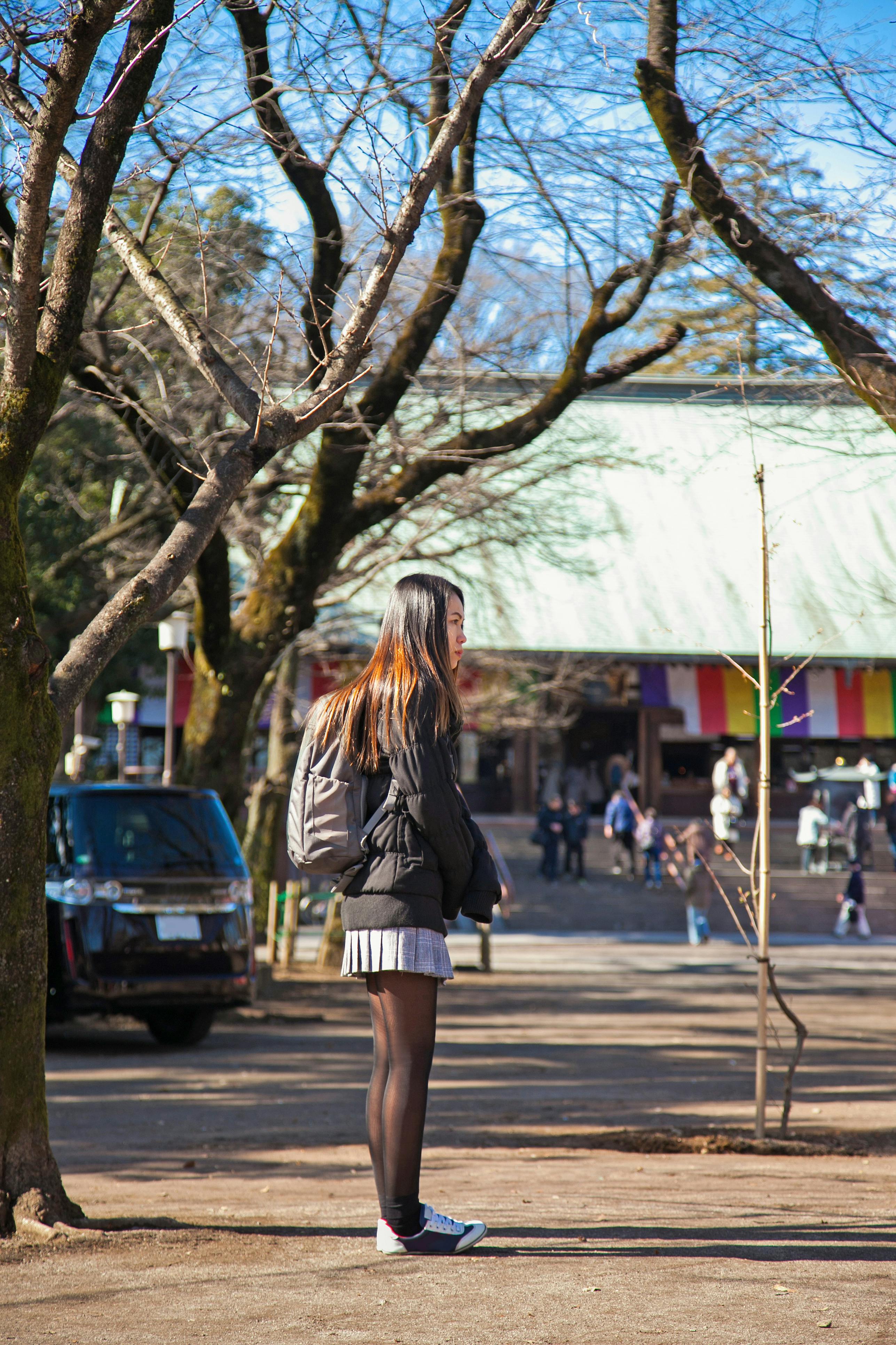 A young woman with long hair, wearing a black jacket, skirt, tights, and a backpack, stands alone in a sunlit park with bare trees and people in the background.