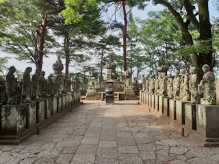 A stone pathway lined with rows of weathered stone statues leads to a central monument, surrounded by lush, green trees in a serene outdoor setting.